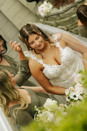 Bride in lace gown and veil smiles while bridesmaids in olive dresses help with white floral bouquet outdoors.