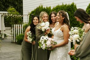 Smiling bride in lace gown poses with four bridesmaids in olive dresses holding white floral bouquets outdoors.