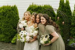 Bride in white lace gown hugging three bridesmaids in olive green dresses holding white floral bouquets outdoors.