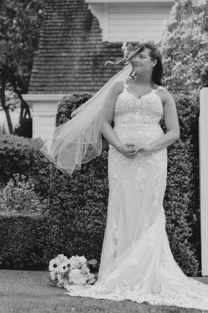 Black and white photo of a bride in lace gown and veil standing outdoors with bouquet.