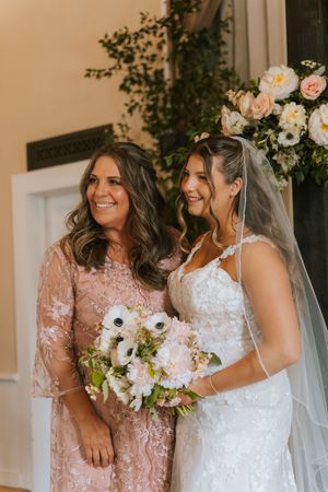 Smiling bride in lace dress holding bouquet stands with woman in pink lace gown at wedding.