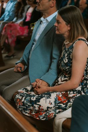 Guests seated in church pews during a wedding ceremony, woman in floral dress beside man in light blue suit.
