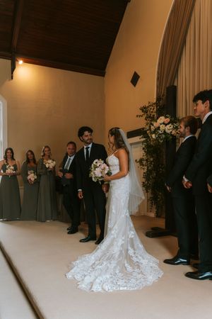 Bride in lace gown and groom in black suit stand at altar during indoor wedding ceremony with bridal party.