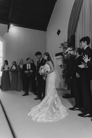 Black and white wedding ceremony photo of bride and groom at altar with bridesmaids and groomsmen in elegant chapel.