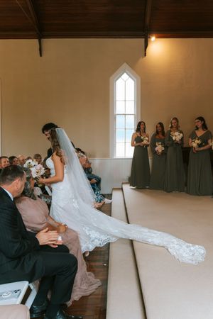 Bride in lace wedding gown with cathedral veil walks down chapel aisle past bridesmaids in sage green dresses.
