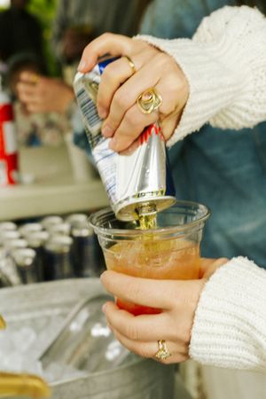 Woman pouring a canned drink into a plastic cup filled with ice at an outdoor event.