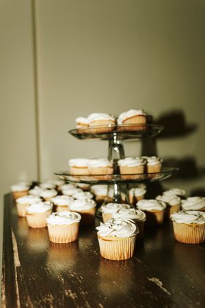 Vanilla cupcakes with white frosting displayed on a tiered glass stand and dark wooden table at a wedding reception.