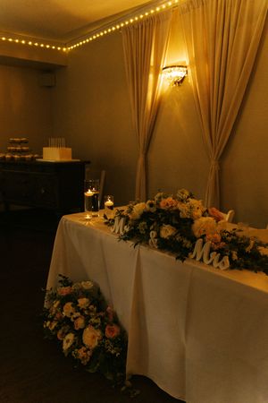 Elegant wedding head table with floral arrangements, Mr & Mrs signs, candles, and draped curtains in warm romantic lighting.