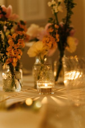 Elegant wedding table centerpiece with glowing candle, glass vases, and colorful orange and pink flowers in warm candlelight.