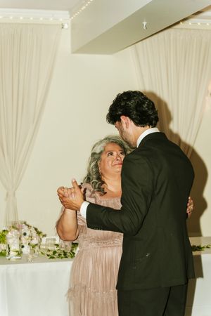 Groom dancing with his mother in elegant wedding reception venue with white draping backdrop.