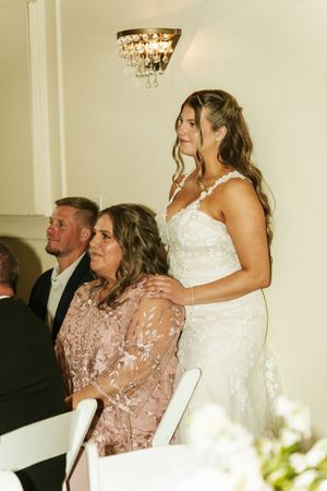 Bride in white lace gown stands beside seated woman in pink floral dress at elegant wedding reception.