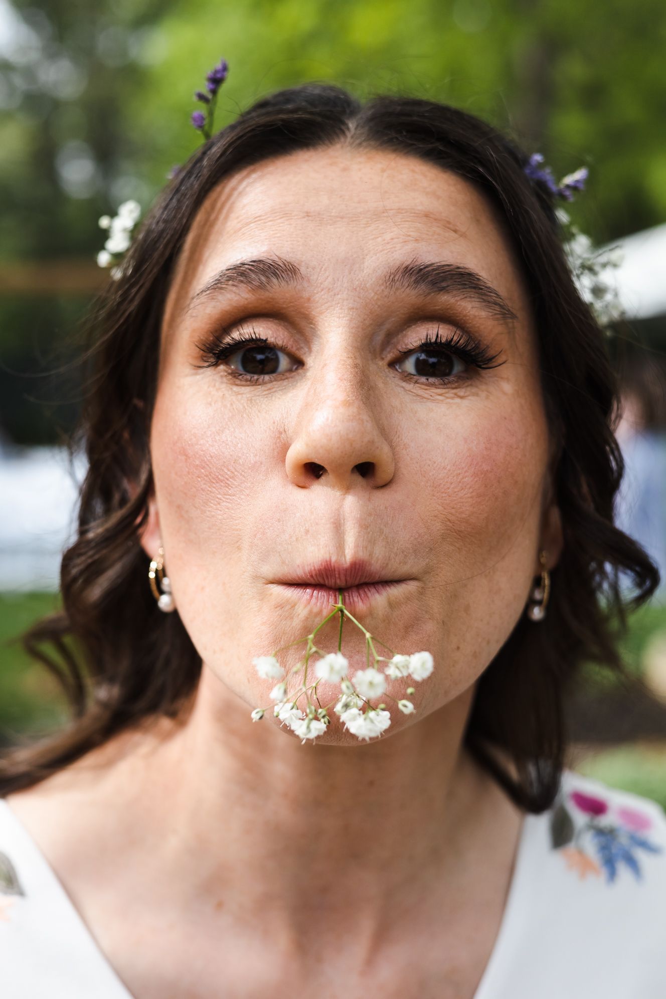 A bride playfully holds white baby's breath flowers in her mouth, wearing pearl earrings and floral hair accessories.