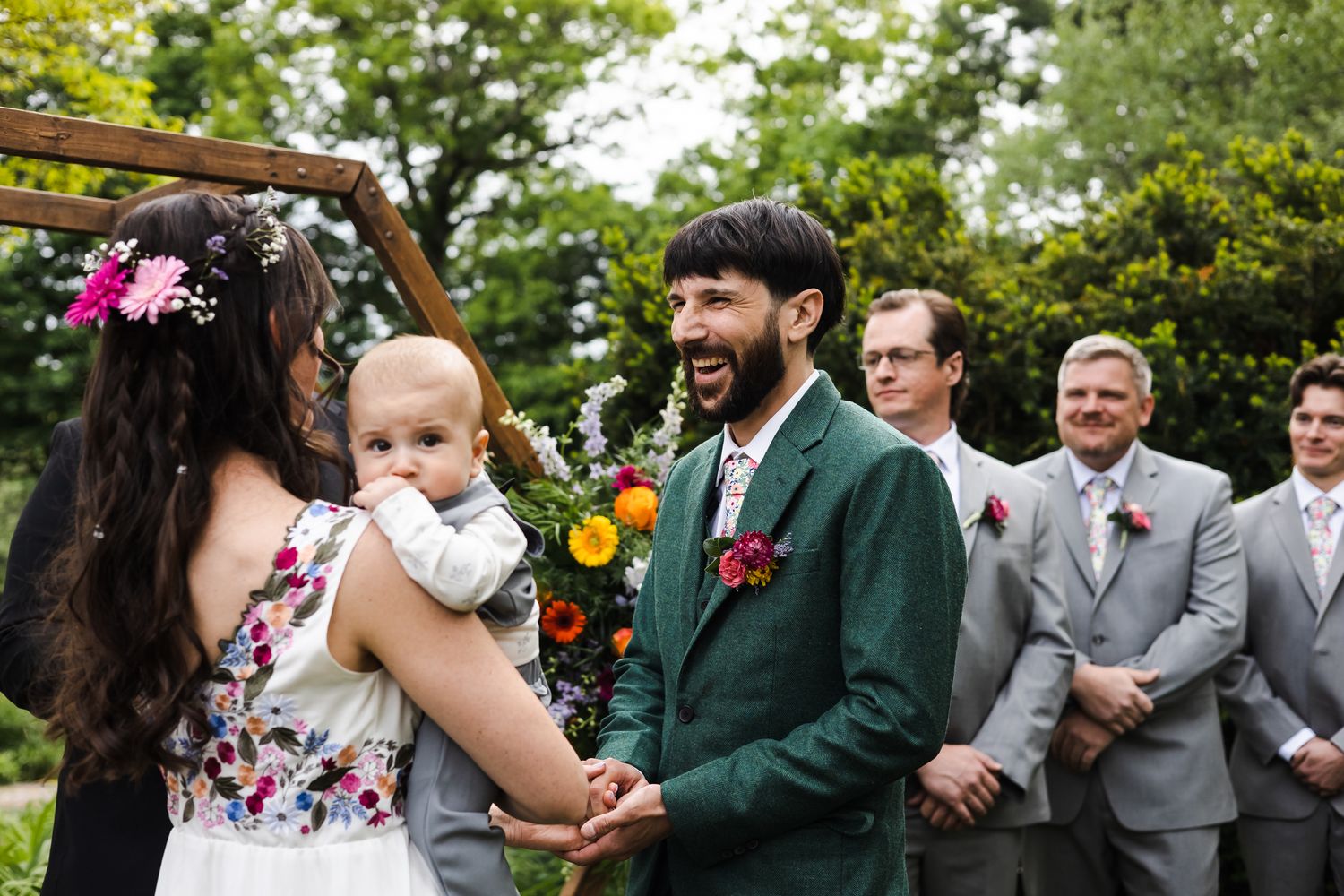 Groom in green suit laughing joyfully with bride holding baby during outdoor wedding ceremony.