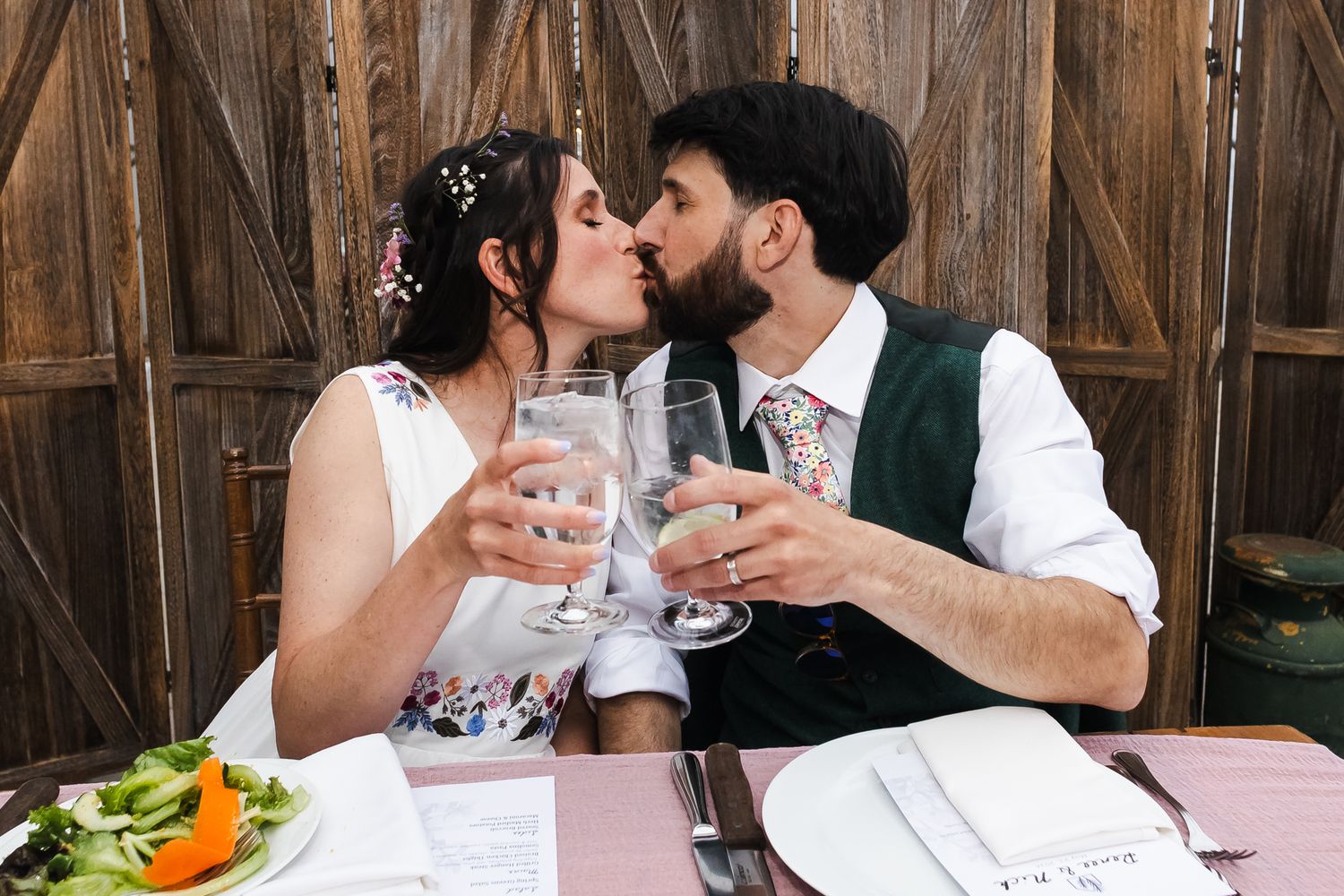 Newlywed couple kissing and clinking wine glasses at wedding reception dinner table in rustic barn venue.