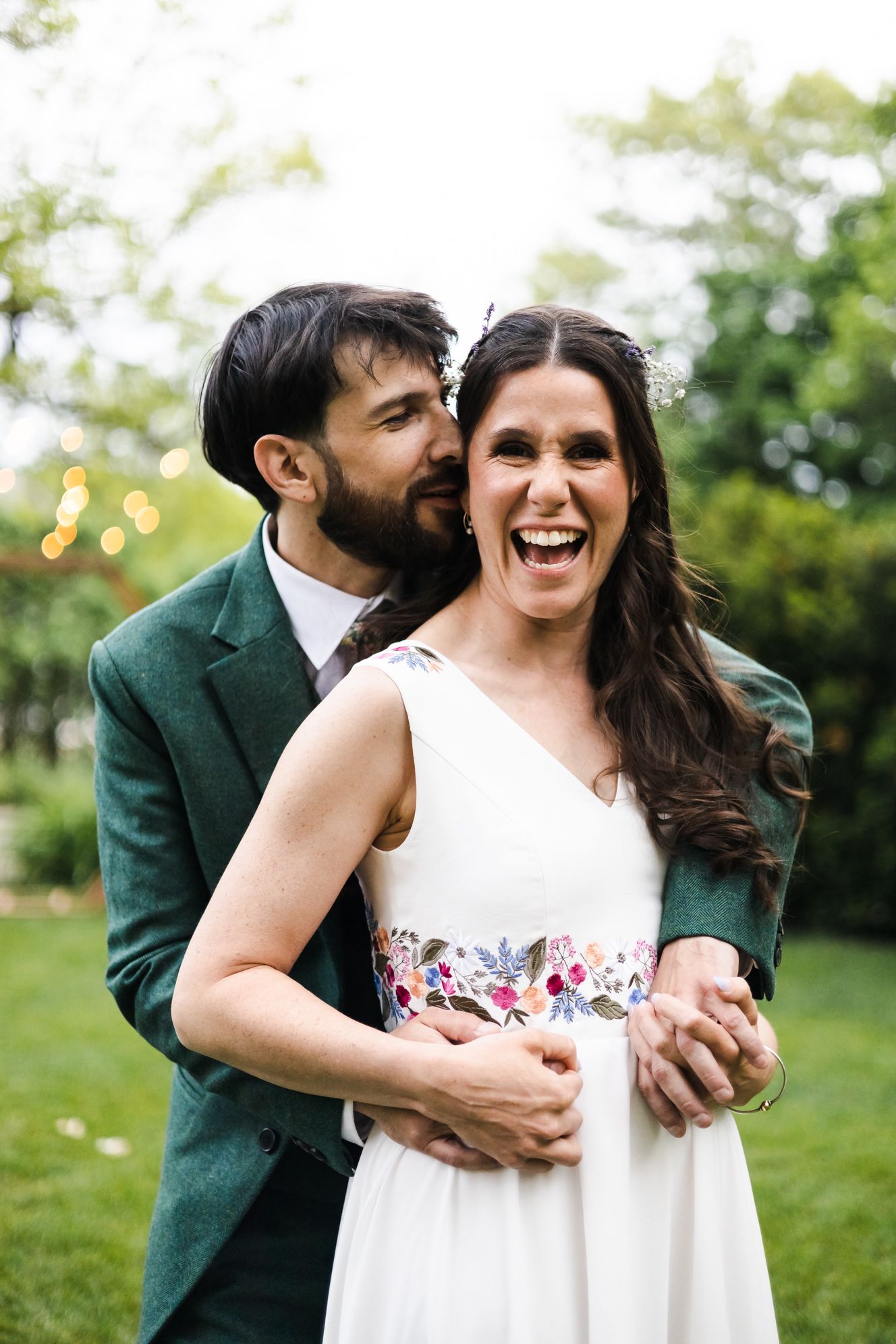 A joyful bride in a white floral-sashed gown laughs as her groom in a green suit embraces her from behind outdoors.