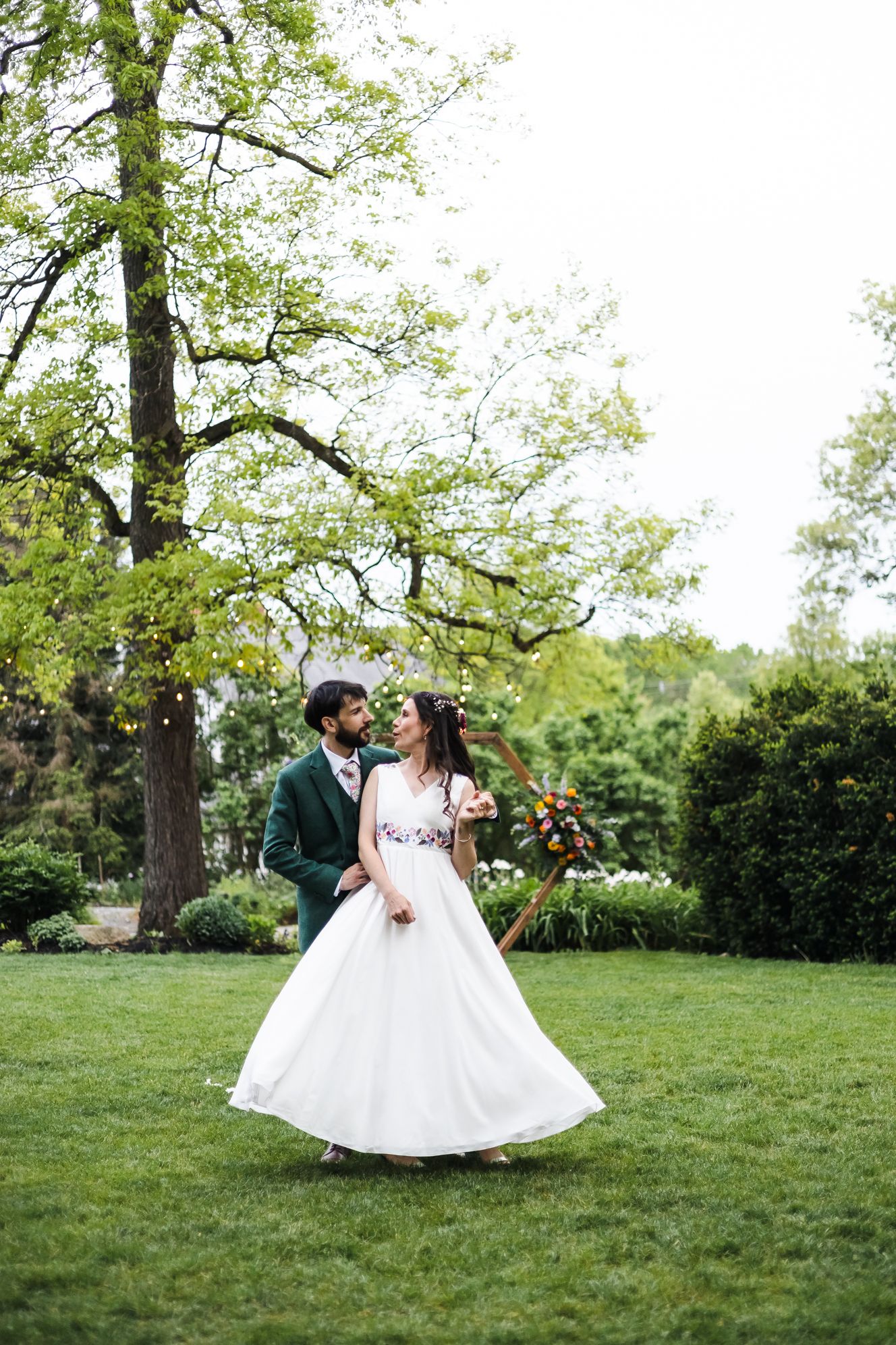 Bride in white gown and groom in green suit embrace on lush garden lawn with trees during outdoor wedding.