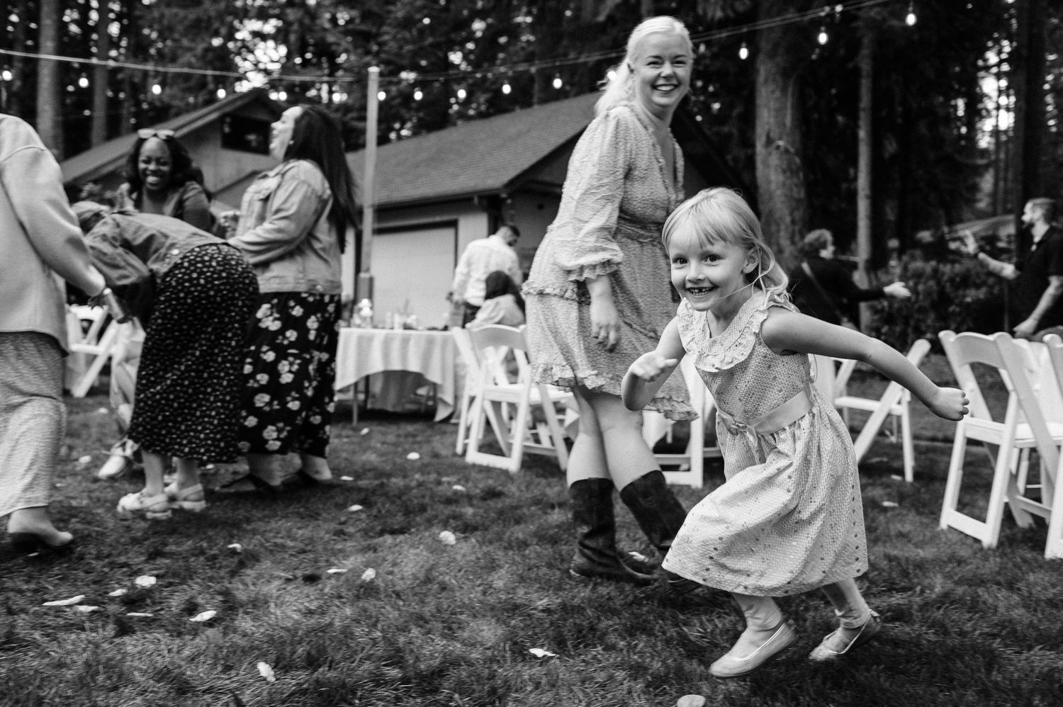 A joyful little girl in a dress runs and dances at an outdoor wedding reception surrounded by guests and white chairs.