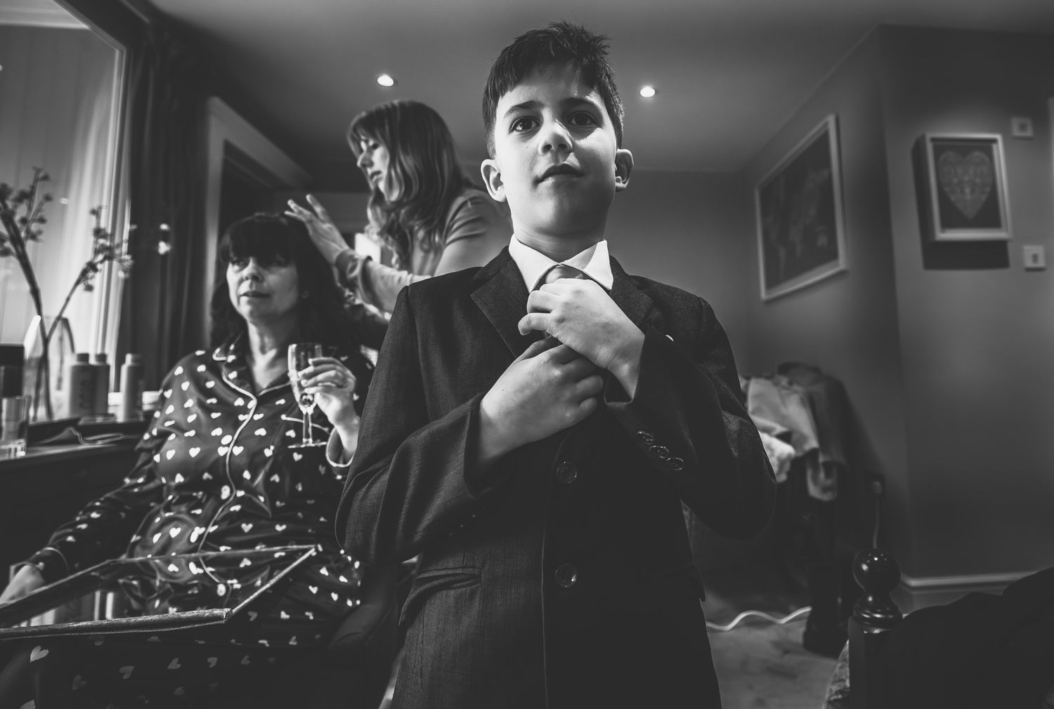 Black and white photo of a young boy in a suit adjusting his tie while women prepare in the background.