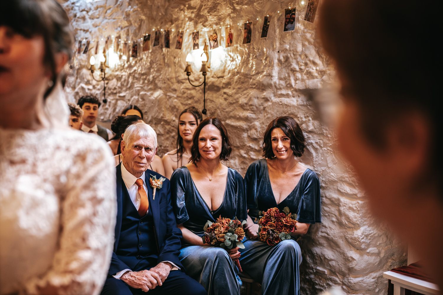 Wedding guests seated in rustic stone venue, bridesmaids in blue velvet dresses holding bouquets, emotional ceremony moment.