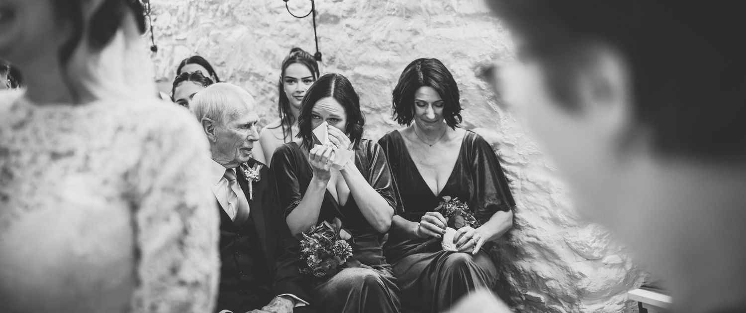 Black and white wedding photo of emotional bridesmaids crying during ceremony in rustic stone venue.