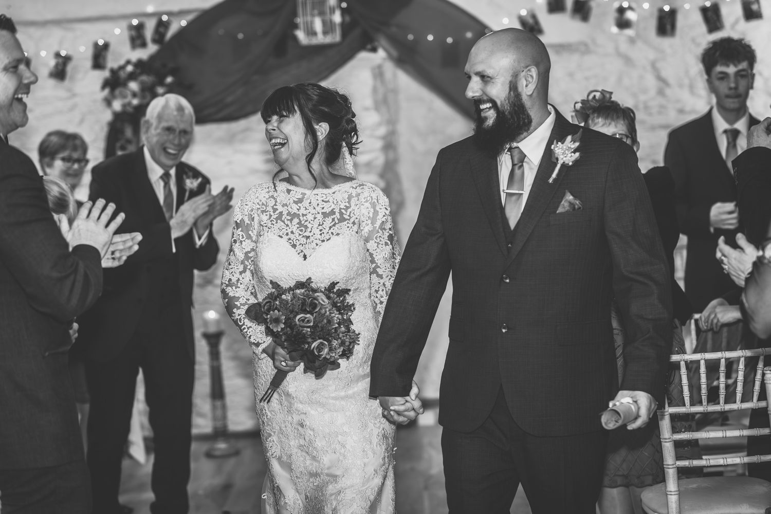 Black and white wedding photo of bride and groom walking down aisle holding hands while guests applaud in decorated venue.