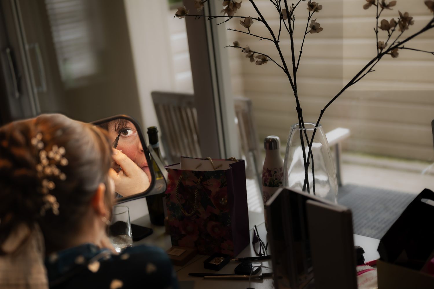 Woman applying makeup at a vanity mirror with dried flowers and beauty accessories on the dresser.