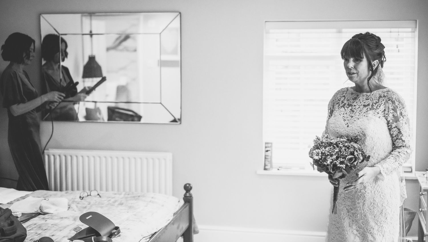 Bride in lace gown holds bouquet while bridesmaid styles hair in bright bedroom before wedding.