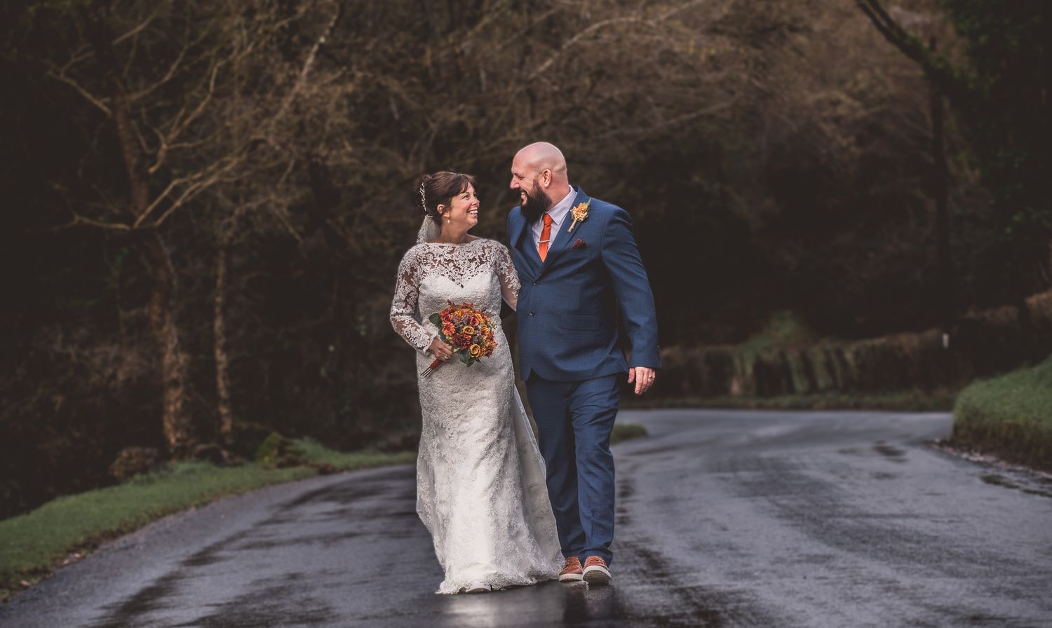 Bride and groom walking hand in hand on a rainy tree-lined road, bride in lace gown, groom in blue suit.