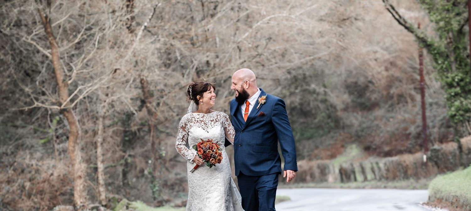 Bride and groom walking hand in hand along a tree-lined path in winter, bride holding an autumn bouquet.