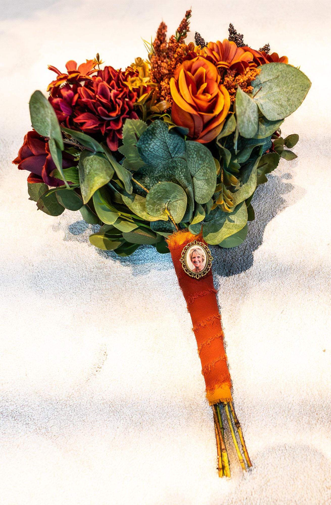 Fall wedding bouquet with orange roses, burgundy dahlias, eucalyptus, and a memorial photo charm on an orange ribbon stem.