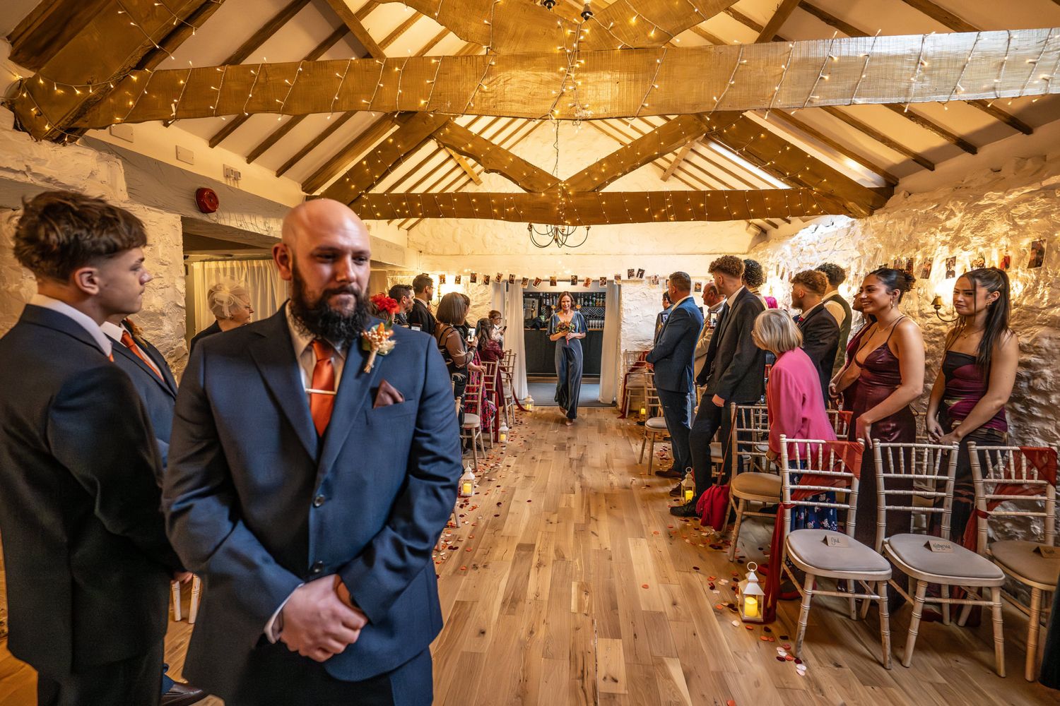 Groom in navy suit with orange tie stands at barn wedding ceremony aisle, guests seated on white chairs.