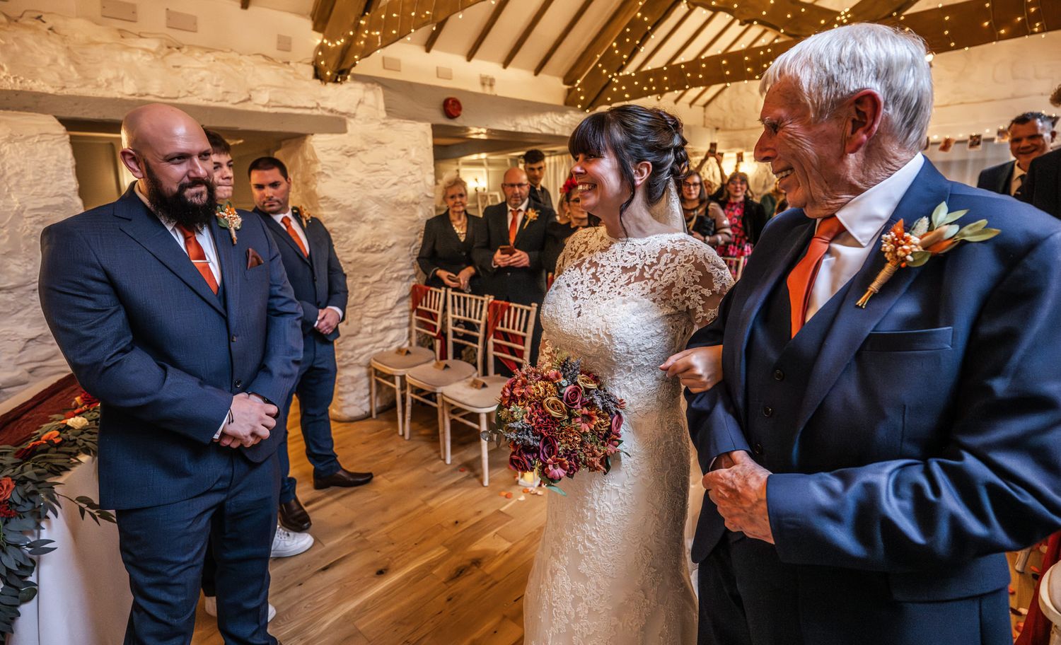 Bride in lace gown meets groom at barn wedding altar, father of bride smiling during ceremony.