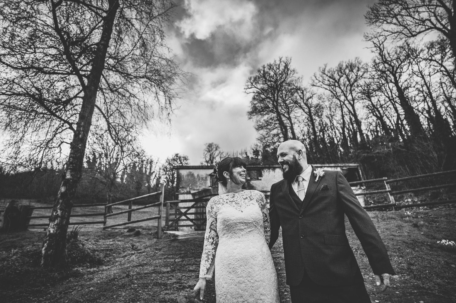 Black and white photo of a bride and groom holding hands outdoors near a rustic fence and bare trees.
