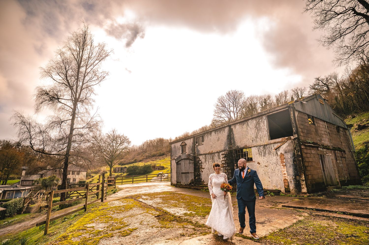 Bride and groom walking hand in hand outside a rustic stone barn venue under dramatic cloudy skies.
