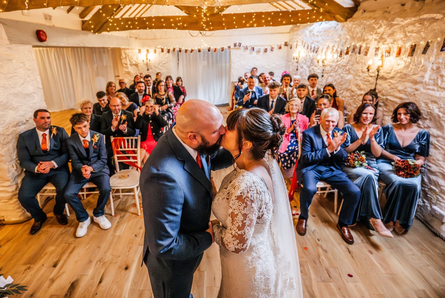 Bride and groom share first kiss at rustic barn wedding ceremony with guests applauding in decorated venue.