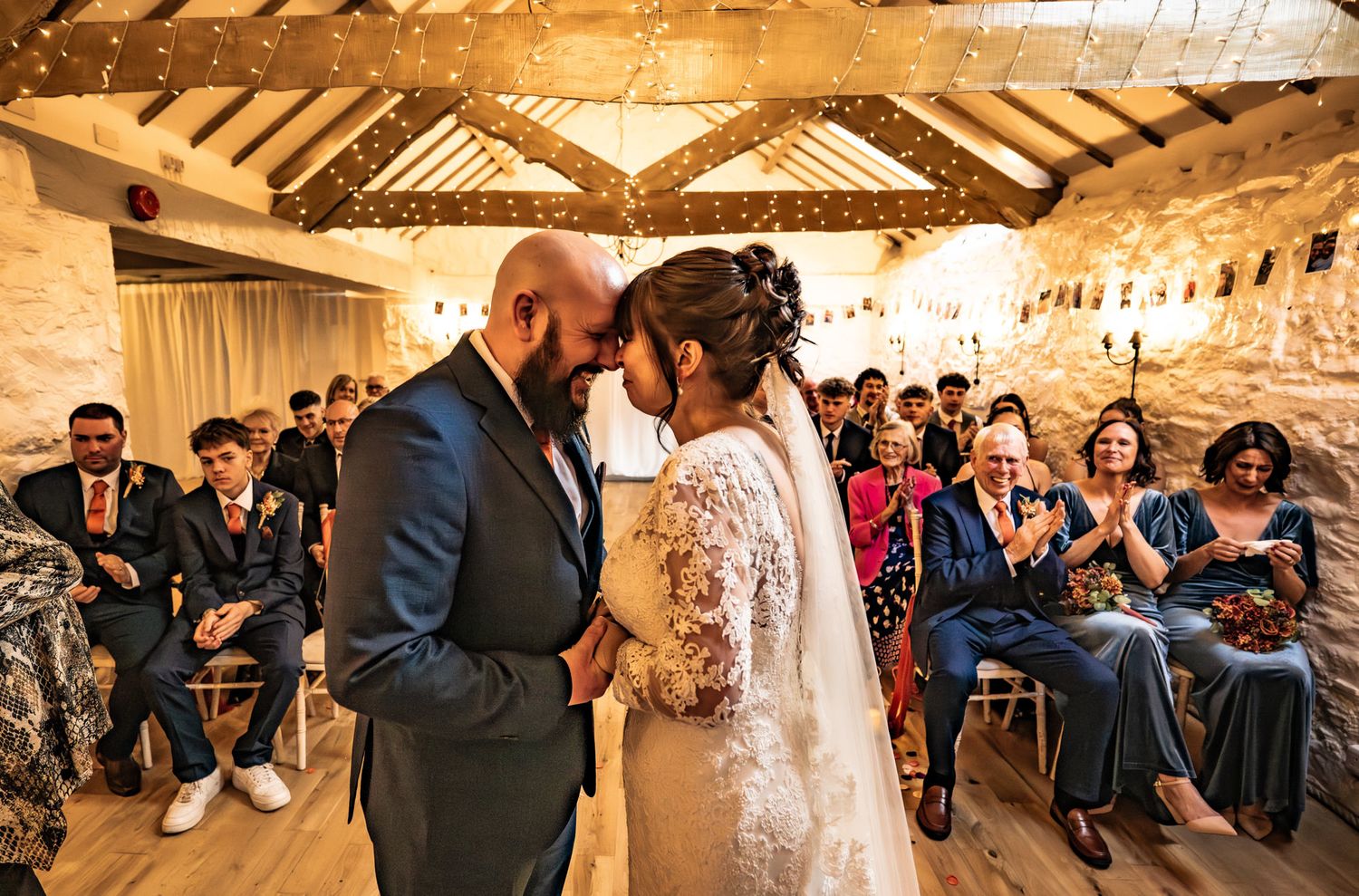 Bride and groom share first kiss at rustic barn wedding ceremony with fairy lights and guests applauding.