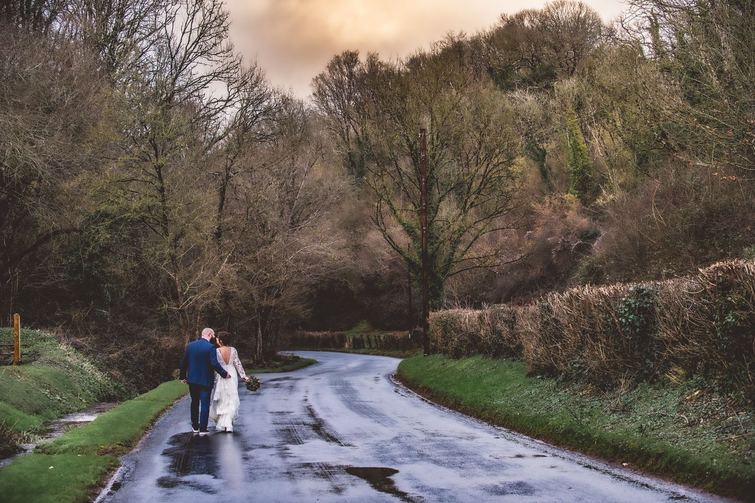 Newlywed couple walking hand in hand down a wet country lane surrounded by autumn trees.