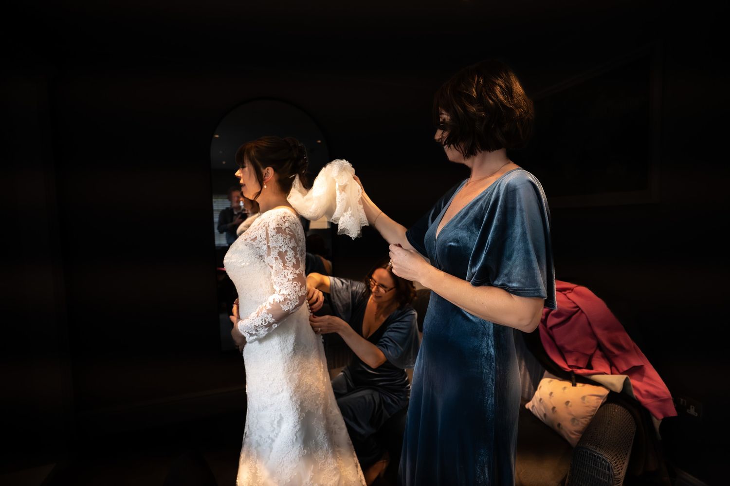 Bridesmaids helping bride into white lace wedding dress backstage in dimly lit venue.
