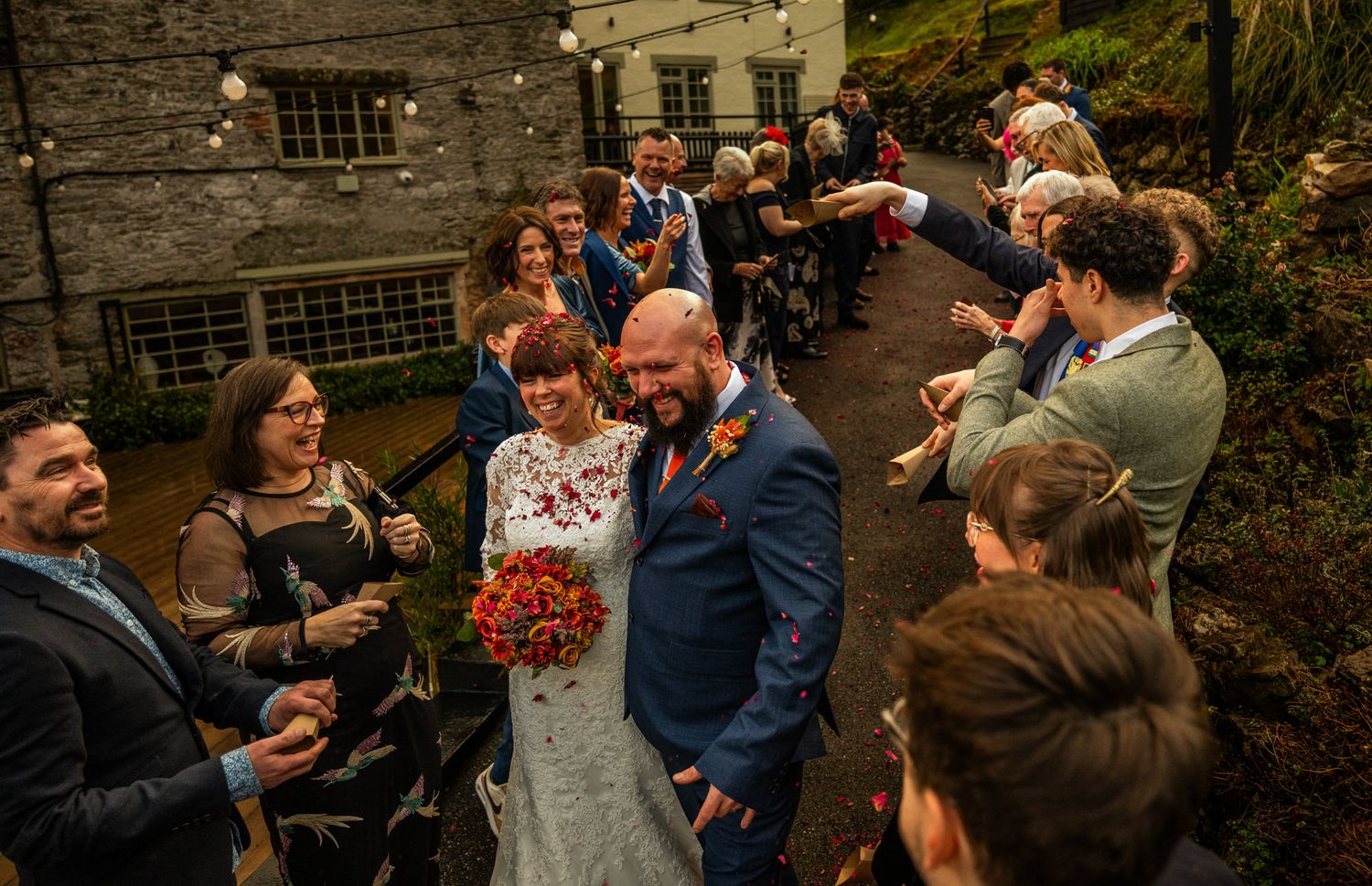 Bride and groom walk through cheering wedding guests outdoors at rustic venue holding red bouquet.
