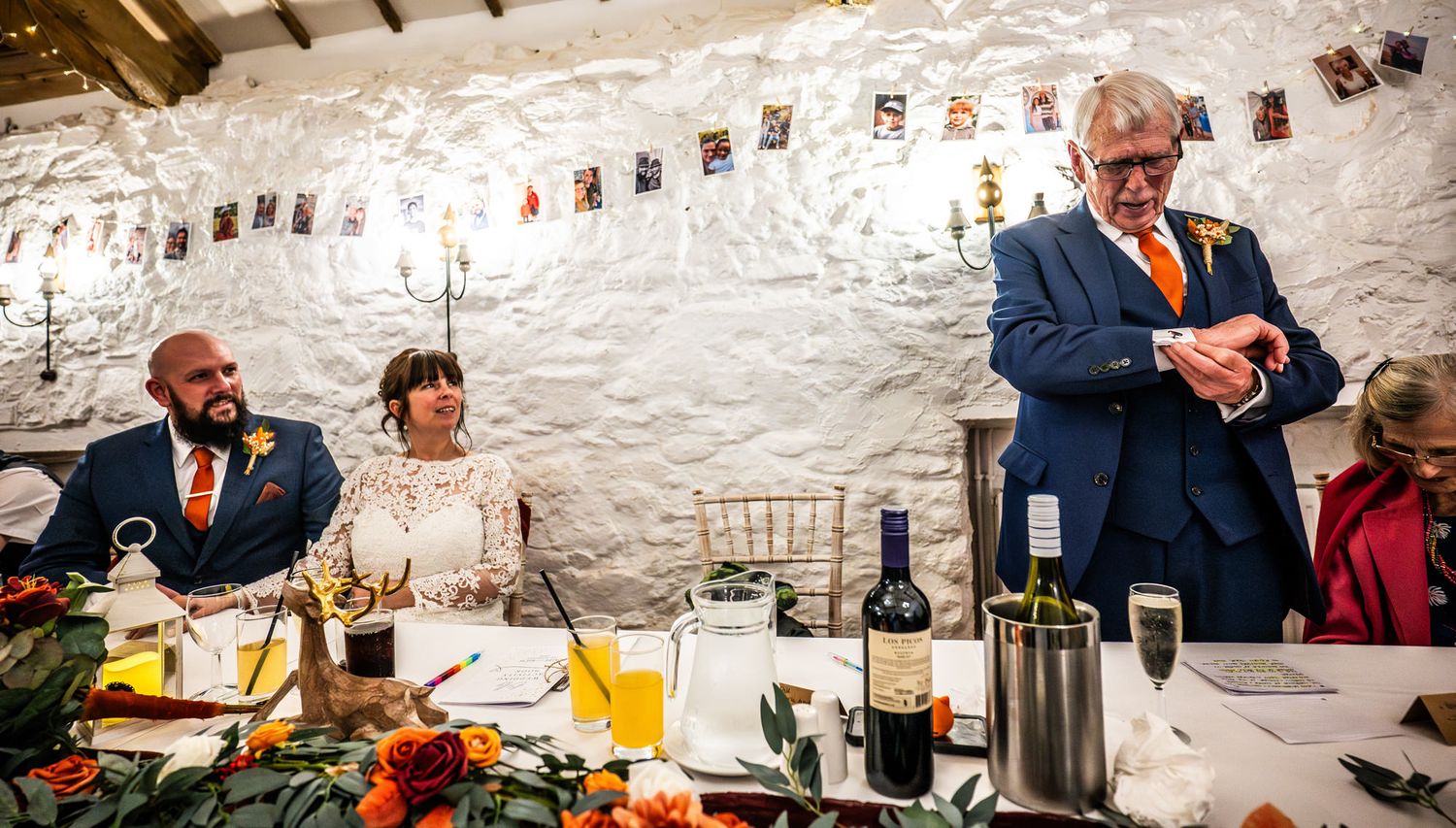 Wedding reception with bride and groom seated at head table while a guest in blue suit adjusts cufflinks in rustic white venue.