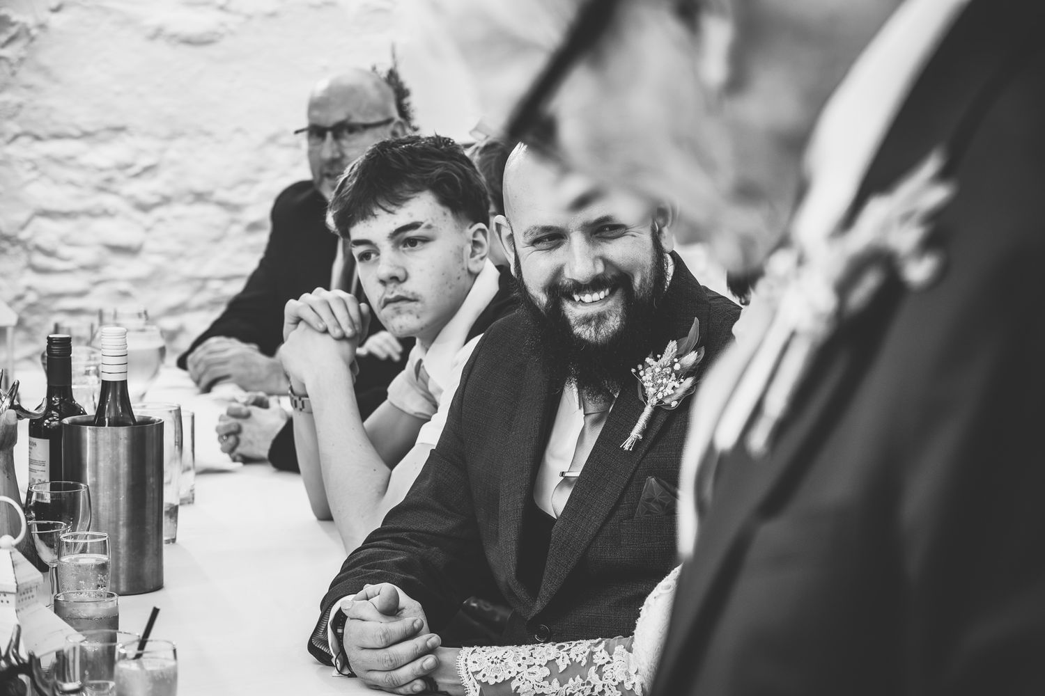 Black and white photo of two smiling men at a wedding reception table, one wearing a boutonniere.