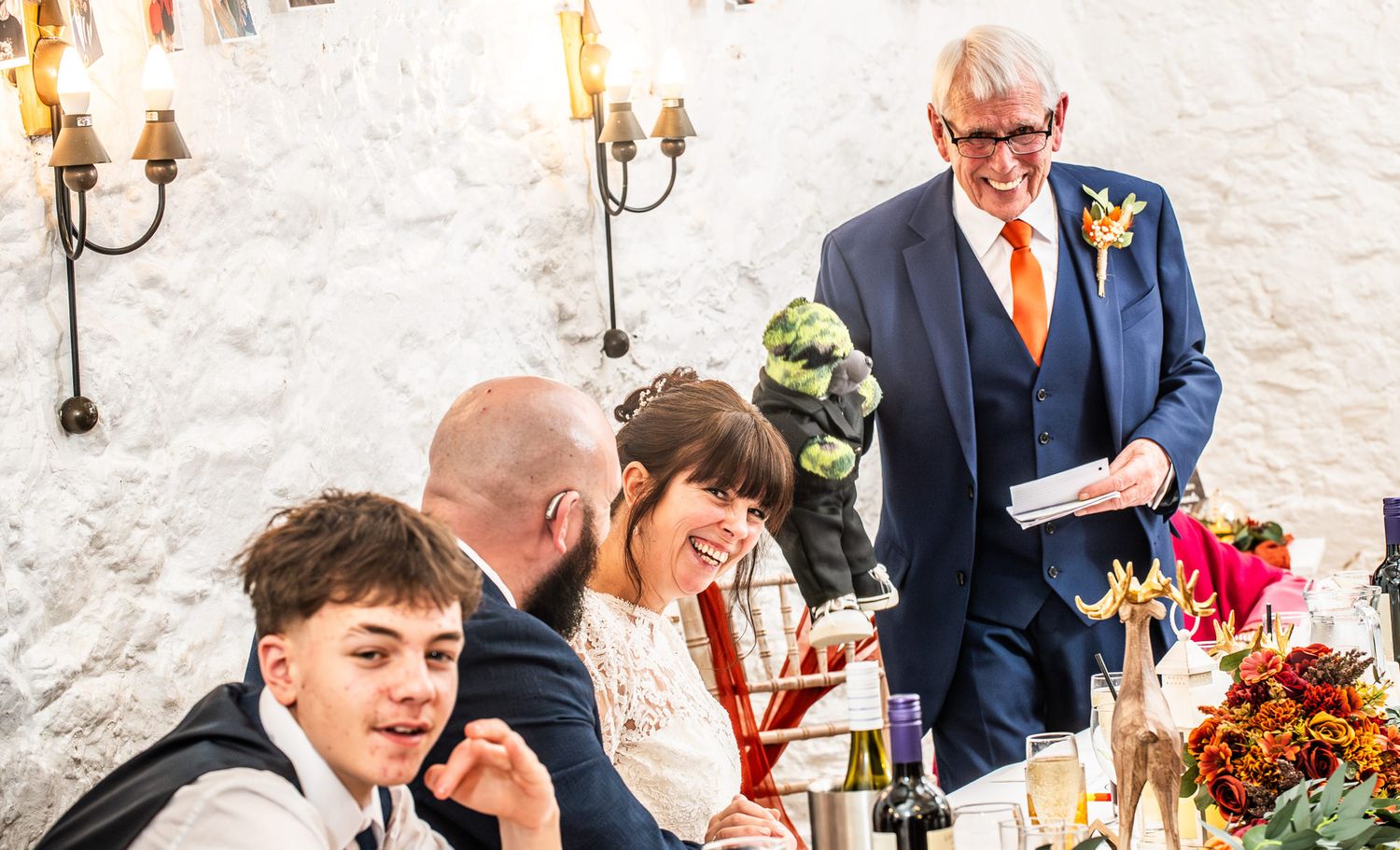 Elderly man in blue suit giving wedding speech to laughing bride and guests at decorated reception table.