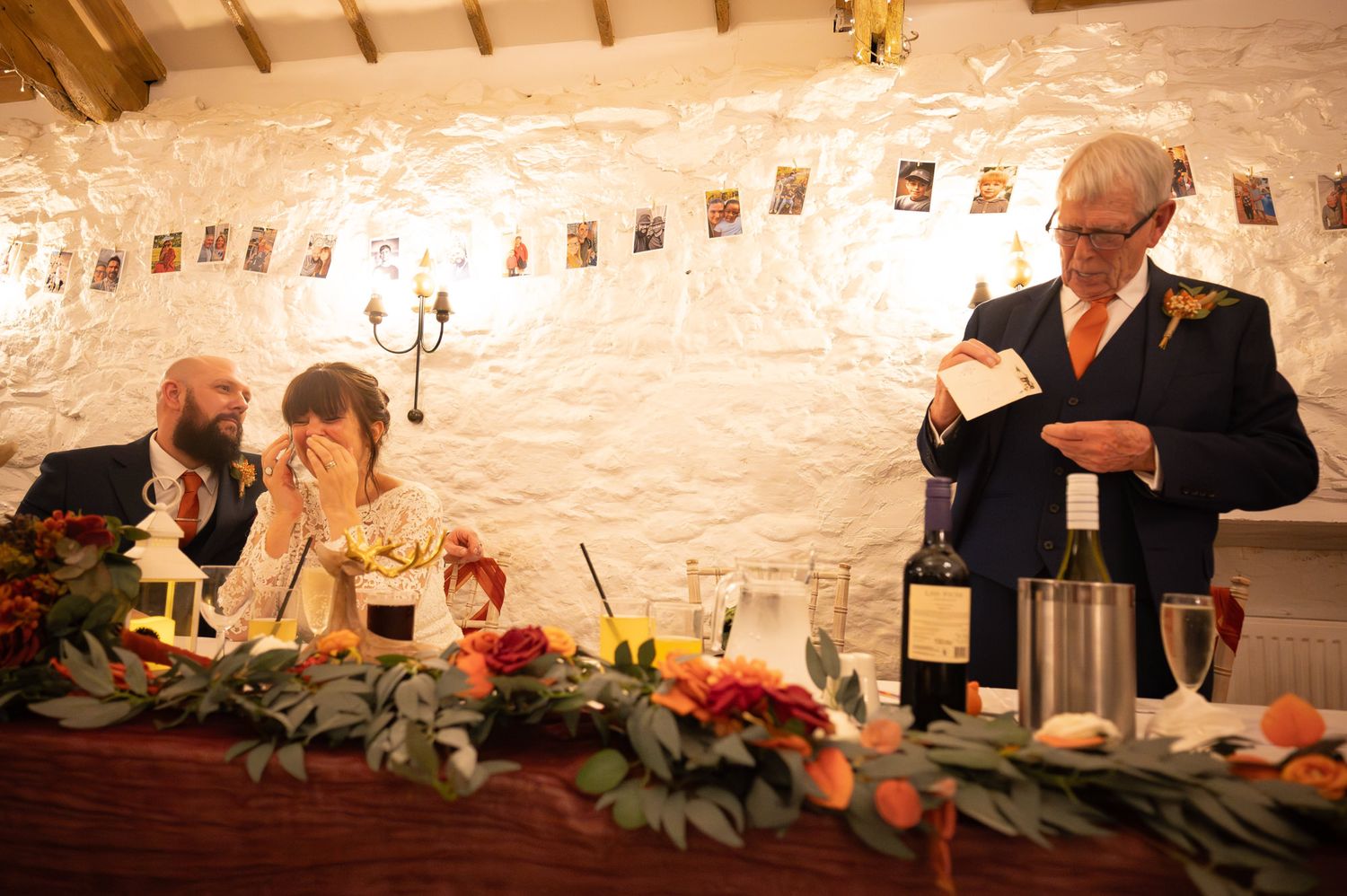 Wedding speaker reads notes while bride covers face laughing at rustic venue with floral table decor.