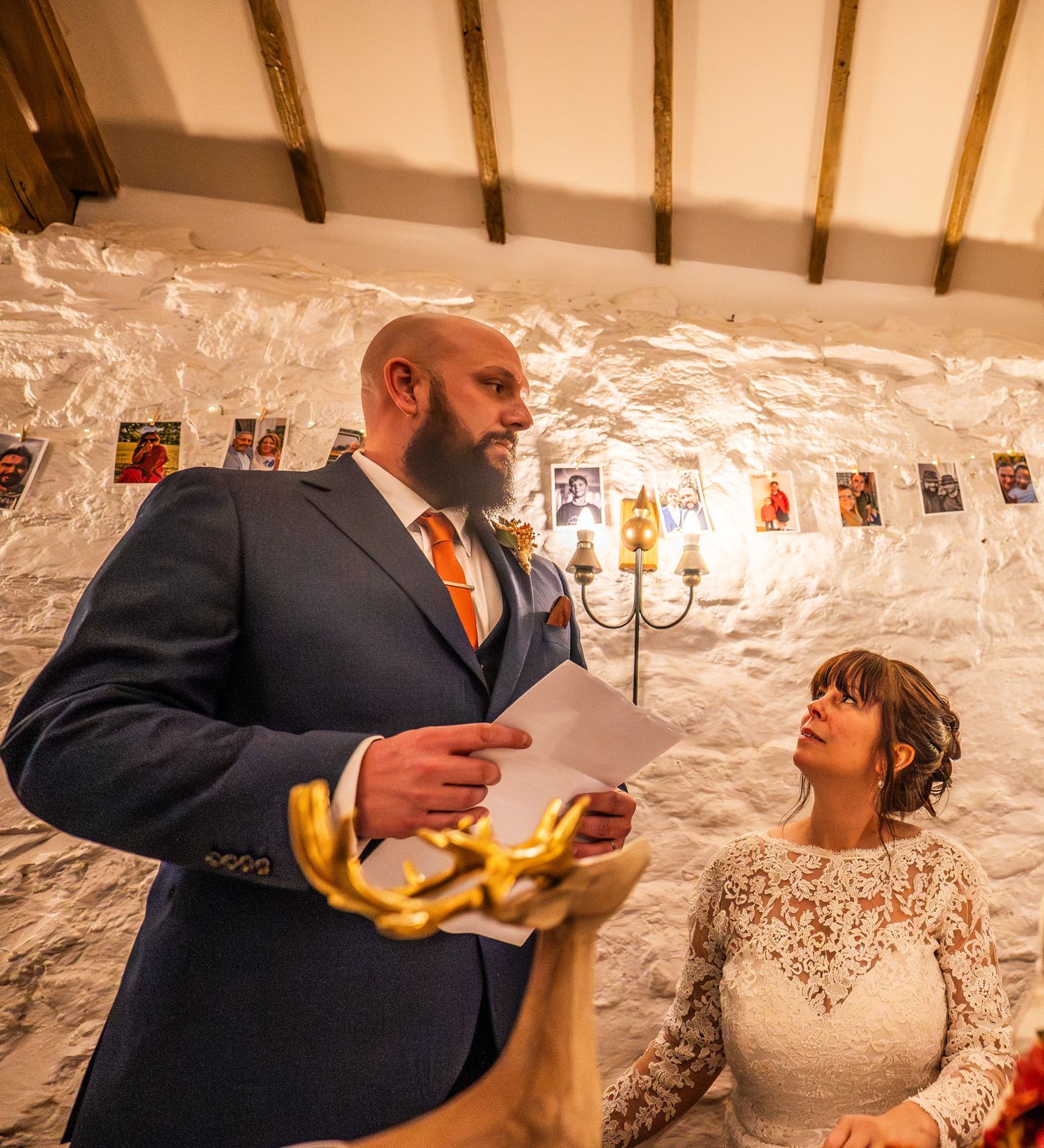 Groom in blue suit reads wedding vows to bride in lace dress during rustic barn ceremony with candlelit stone walls.