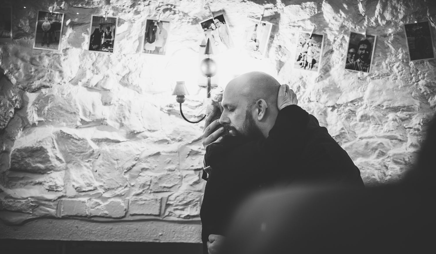 Black and white photo of a bald man in dark clothing sitting pensively in a rustic stone-walled room with wall sconce.