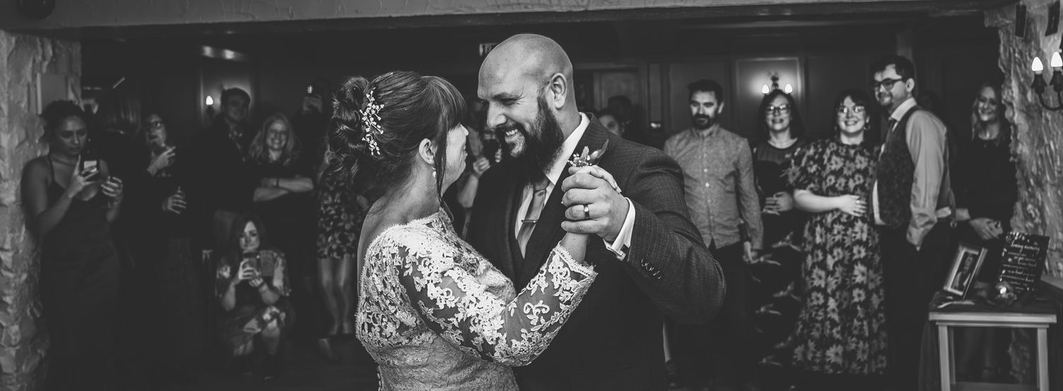 Black and white photo of a couple sharing their first dance at a wedding reception with guests watching in the background.