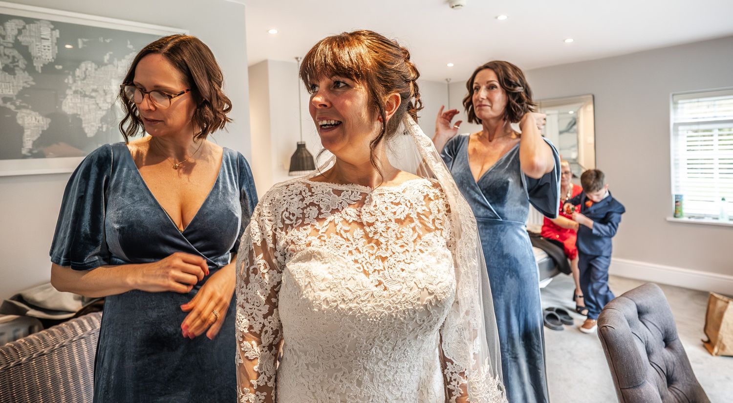 Bride in white lace gown with bridesmaids in blue dresses helping her prepare for wedding ceremony indoors.