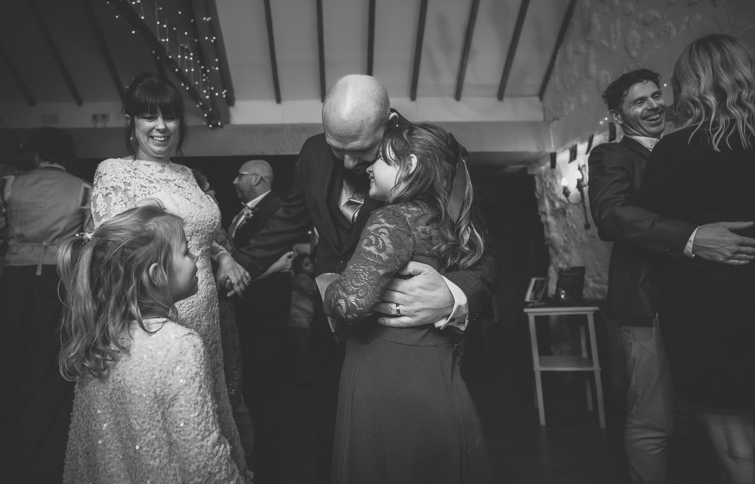Black and white wedding photo of a bald groom embracing a guest on the dance floor surrounded by smiling attendees.