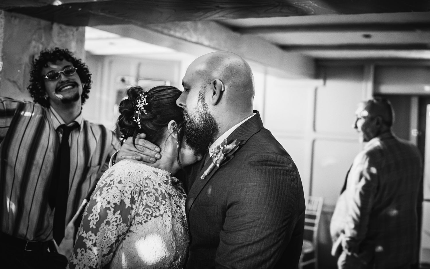 Black and white wedding photo of couple sharing first kiss, guests smiling in background.