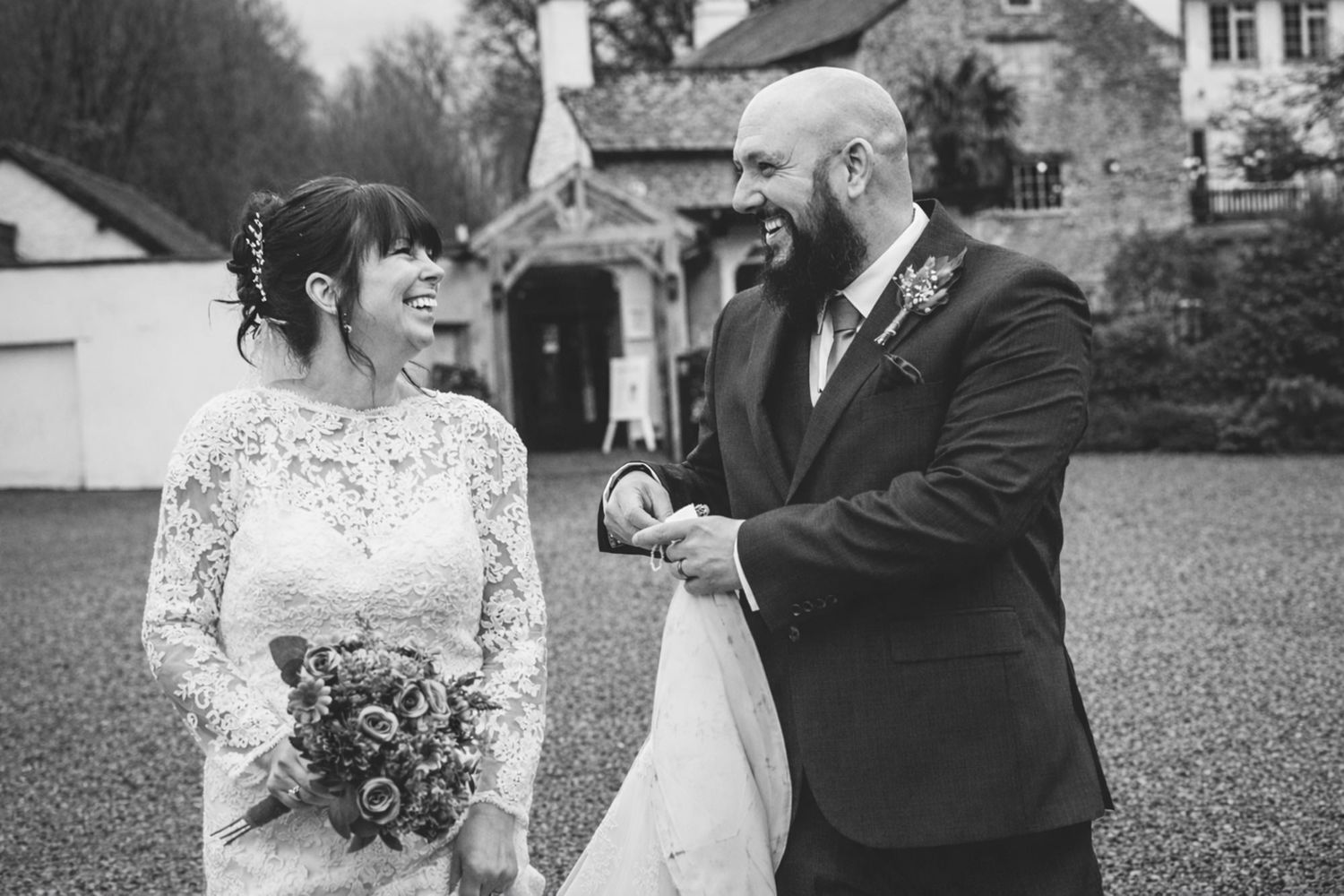 Black and white photo of a smiling bride holding bouquet and groom holding hands outdoors at a wedding venue.